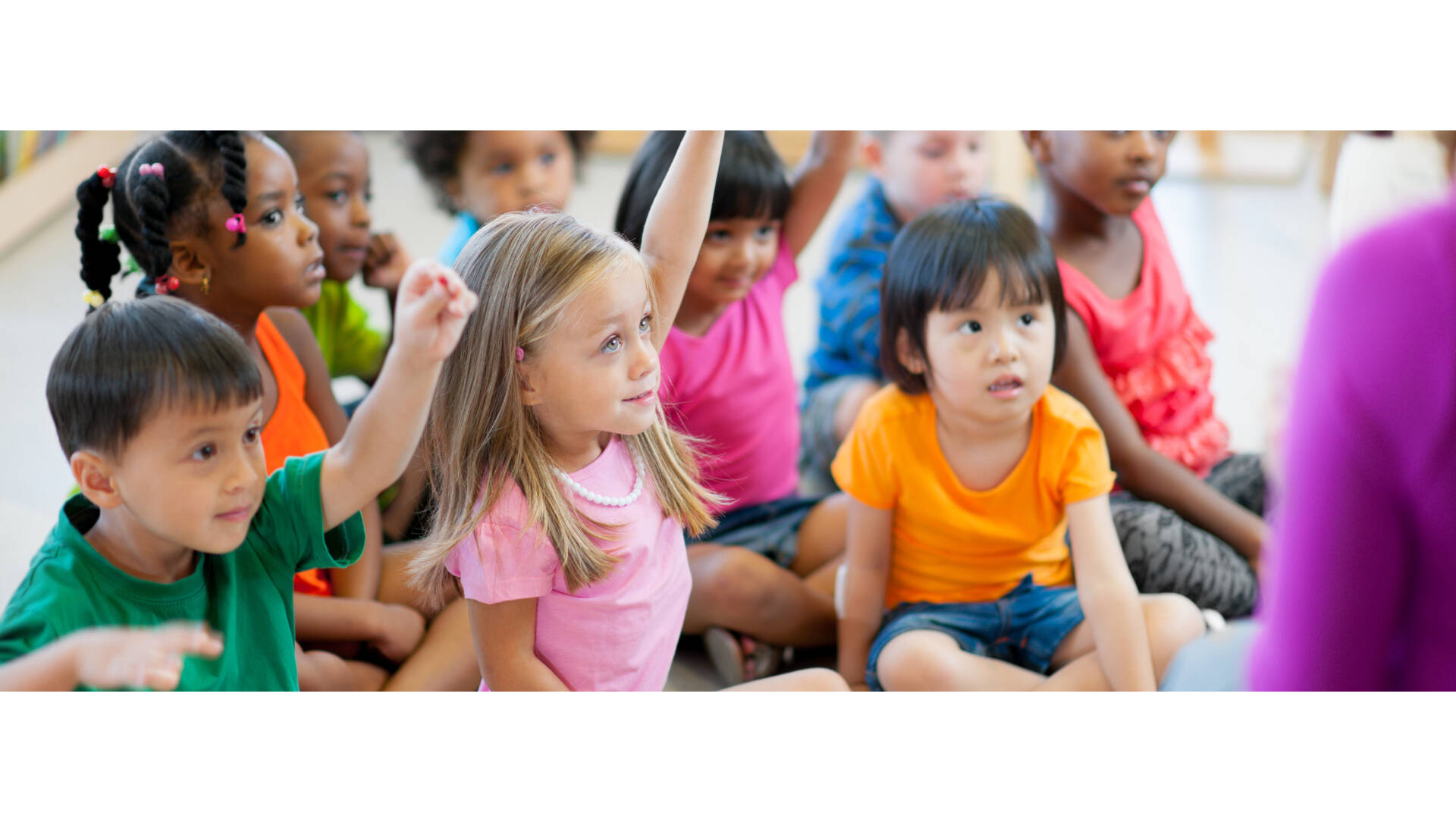 Children in a preschool classroom sitting on the floor listening to their teacher and raising hands.